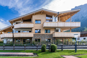 Neubau einer mehrstöckigen Wohnhausanlage mit Holzfassade, großen Balkonen und steinverklehrtem Erdgeschoss vor Bergkulisse.
